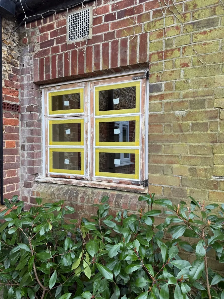 Timber casement window mid-restoration with yellow masking tape and primer in a brick wall